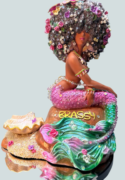 Stunning hand-painted African American mermaid figurine named Lumira from the Brassy collection. Features a lavender and green glittering tail, a pearl-studded pink top, and a voluminous flower-filled afro. Designed by Clover Jené Mermaid.