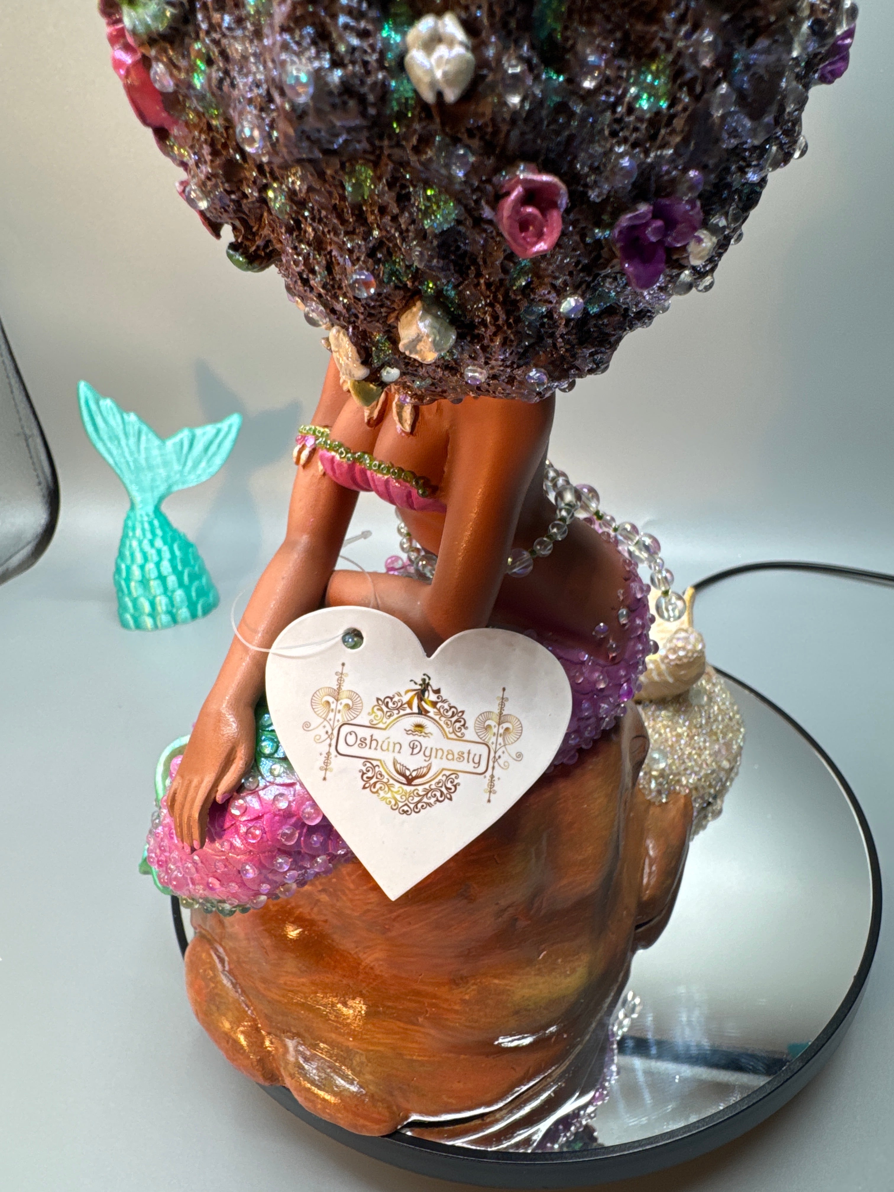 Back view of Lumira’s glossy, hand-painted form revealing her luminous Afro hair, radiant tail, and the crowned Oshún Dynasea tag—marking her as royalty in every sculpted detail.