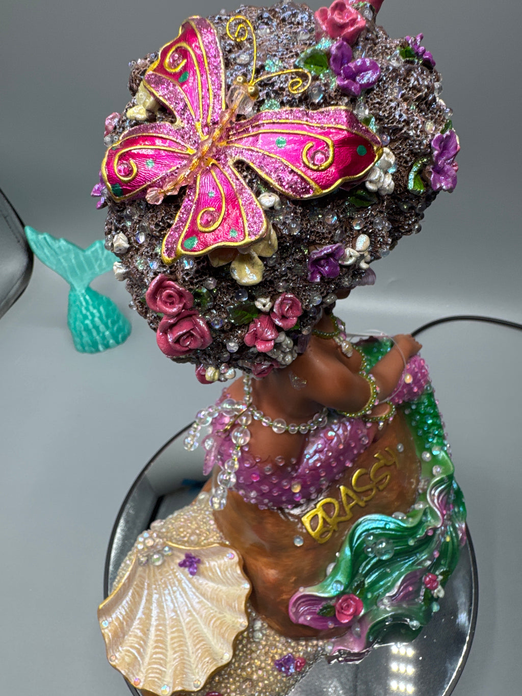 Close-up of Lumira’s floral afro featuring a metallic pink butterfly, iridescent bubbles, and glossy luminescent accents. A radiant handcrafted Black mermaid figurine from the Brassy collection.
