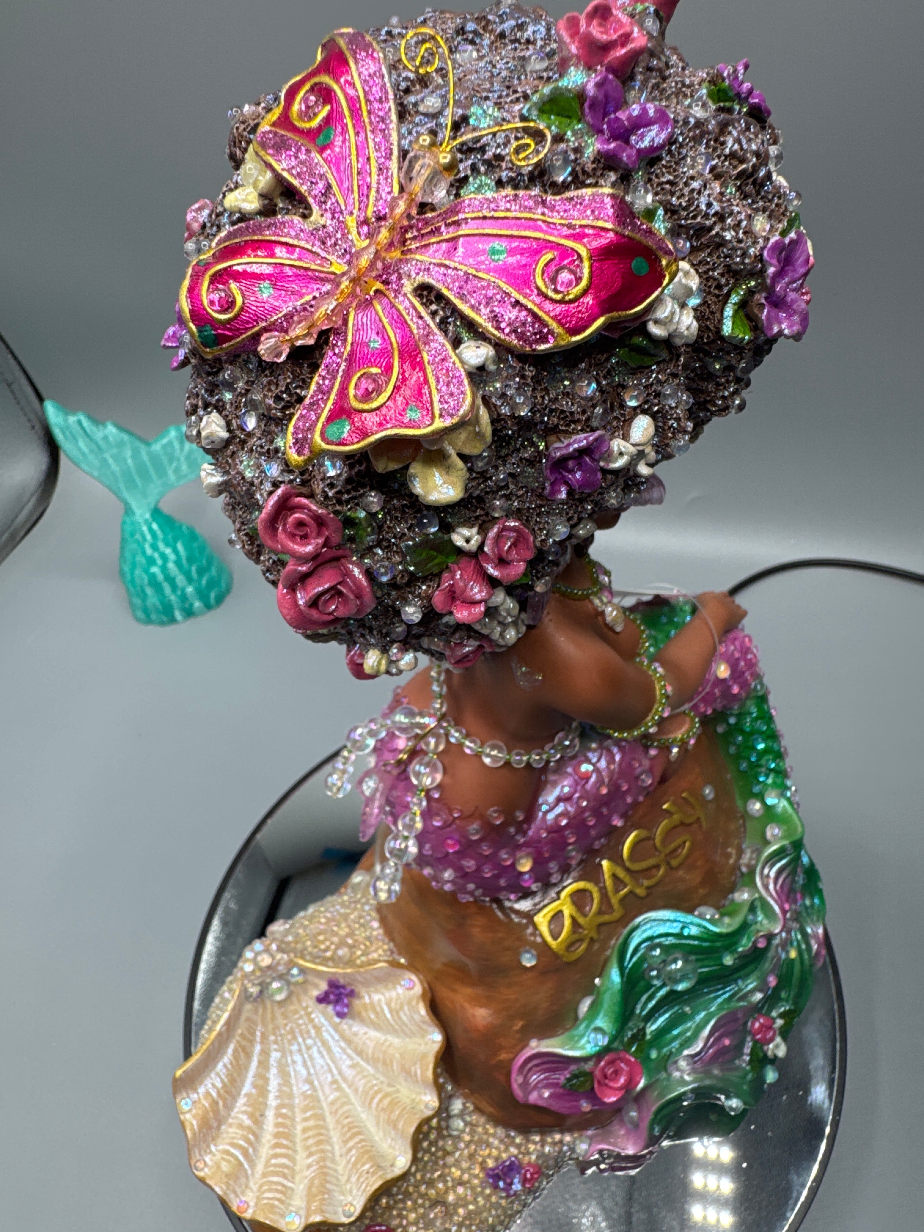 Close-up of Lumira’s floral afro featuring a metallic pink butterfly, iridescent bubbles, and glossy luminescent accents. A radiant handcrafted Black mermaid figurine from the Brassy collection.
