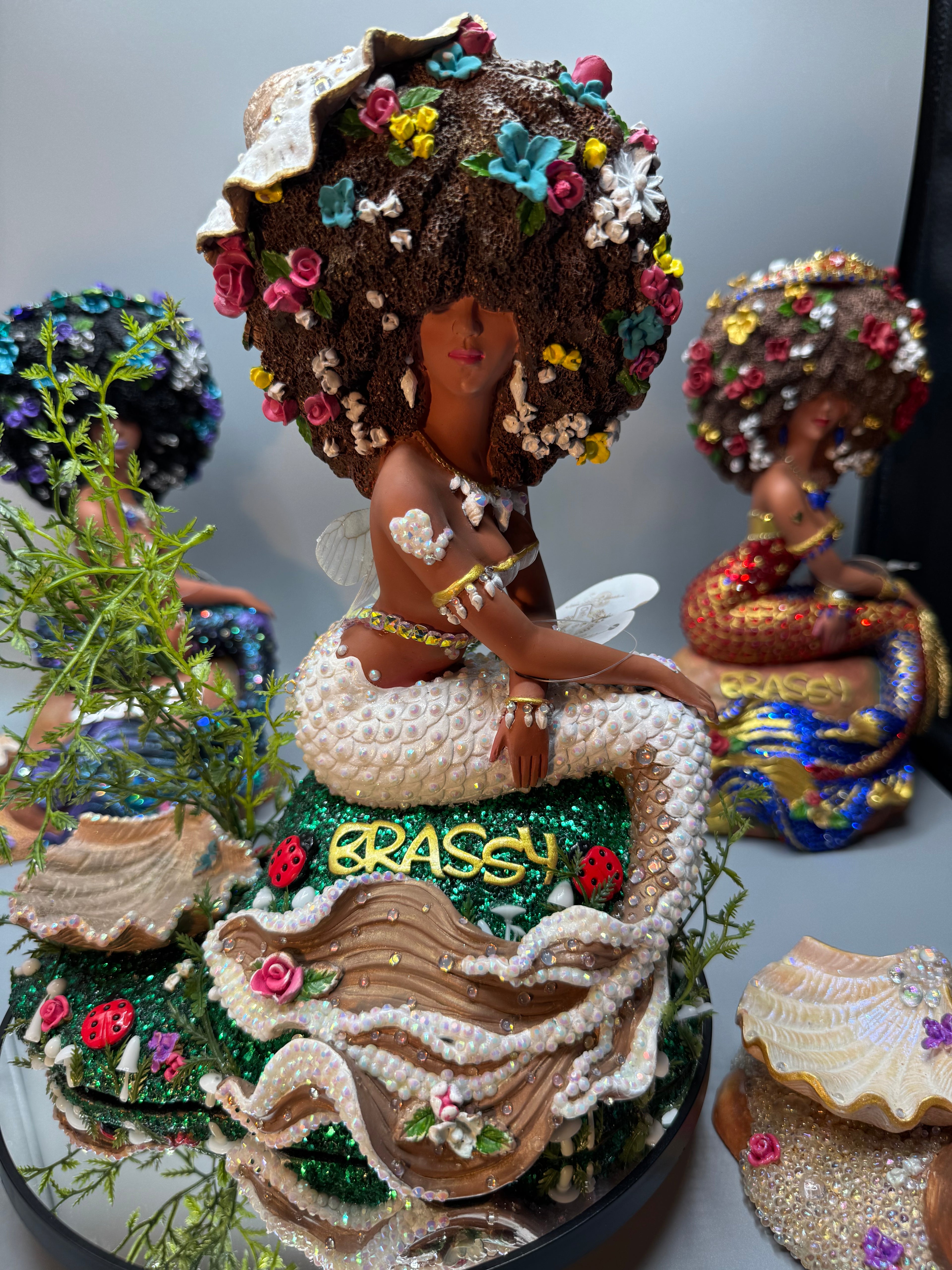 Afro Fairy Mermaid statue with mushroom hat and glitter grass base. Hand-painted OOAK fantasy décor from the Clover Unleashed collection. Features rhinestone-covered white and brown tail, green glitter base with mushroom clusters, and bold Afro hairstyle. Black mermaid art by Clover Jené Mermaid.